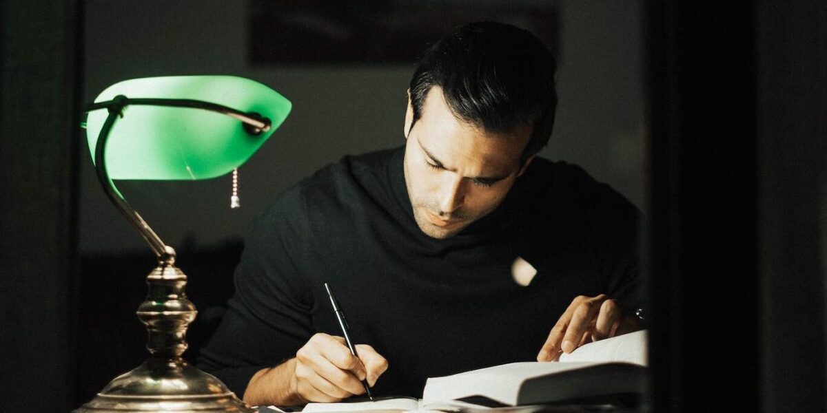 Concentrated young male wearing black turtleneck taking notes and reading books while sitting at desk with papers and bankers lamp in dark home office